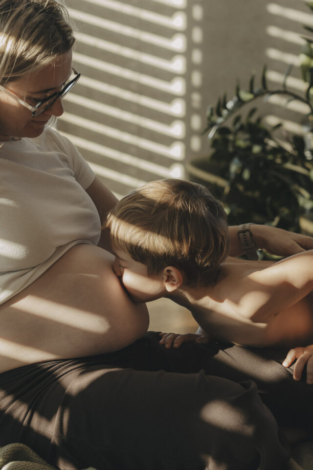 Junge, der Babybauch seiner Mama küsst.