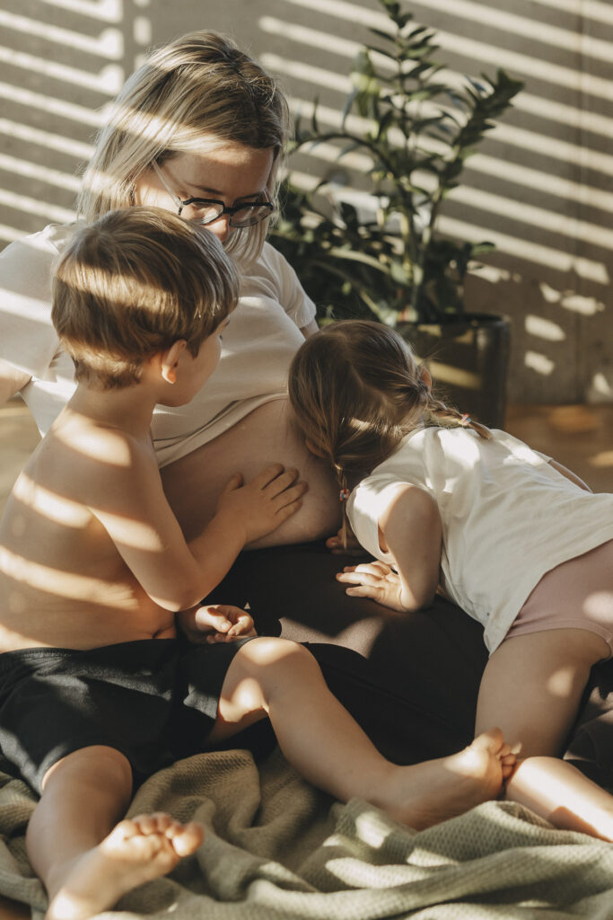 Mama mit Babybauch und den zwei Geschwisterkindern, die den Bauch streicheln und hineinhorchen.