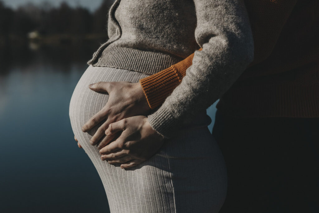 Eine Frau in einem beigen Kleid. Man sieht den Ausschnitt von der Brust bis zur Hüfte, aus der Seitenansicht. Mit den hälten erfasst sie die Hände ihres Mannes, der dicht hinter ihr steht und behutsam ihren Babybauch hält.