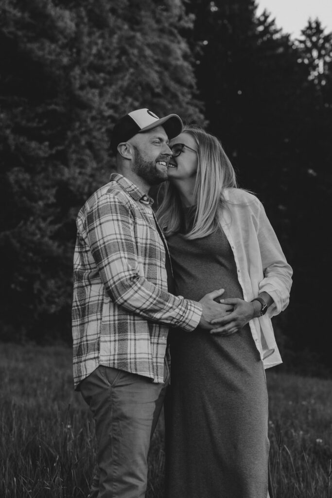 Schwarz-Weiß-Bild. Eine Frau küsst den Mann auf die Wange, er lächelt in die Ferne und berührt sanft ihren Babybauch.