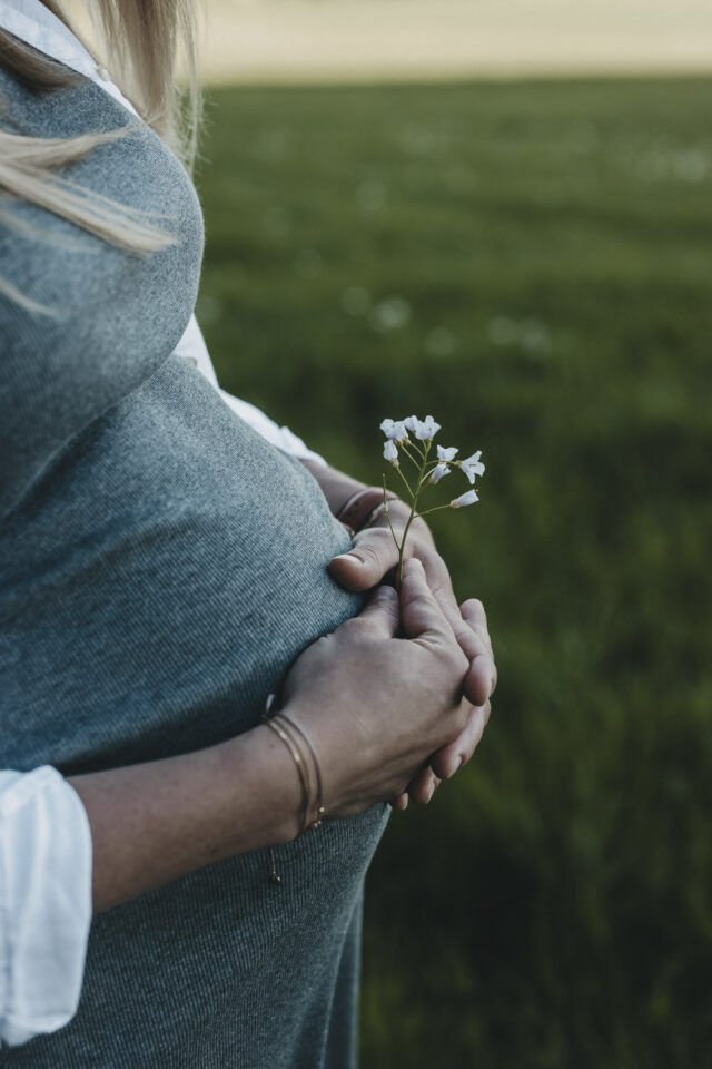 Eine blonde Frau in einem grauen Kleid. Man sieht den Ausschnitt von der Brust bis zur Hüfte, aus der Seitenansicht. Die Hände hat sie vor ihrem Bauch verschränkt und hält eine weiße Blume.