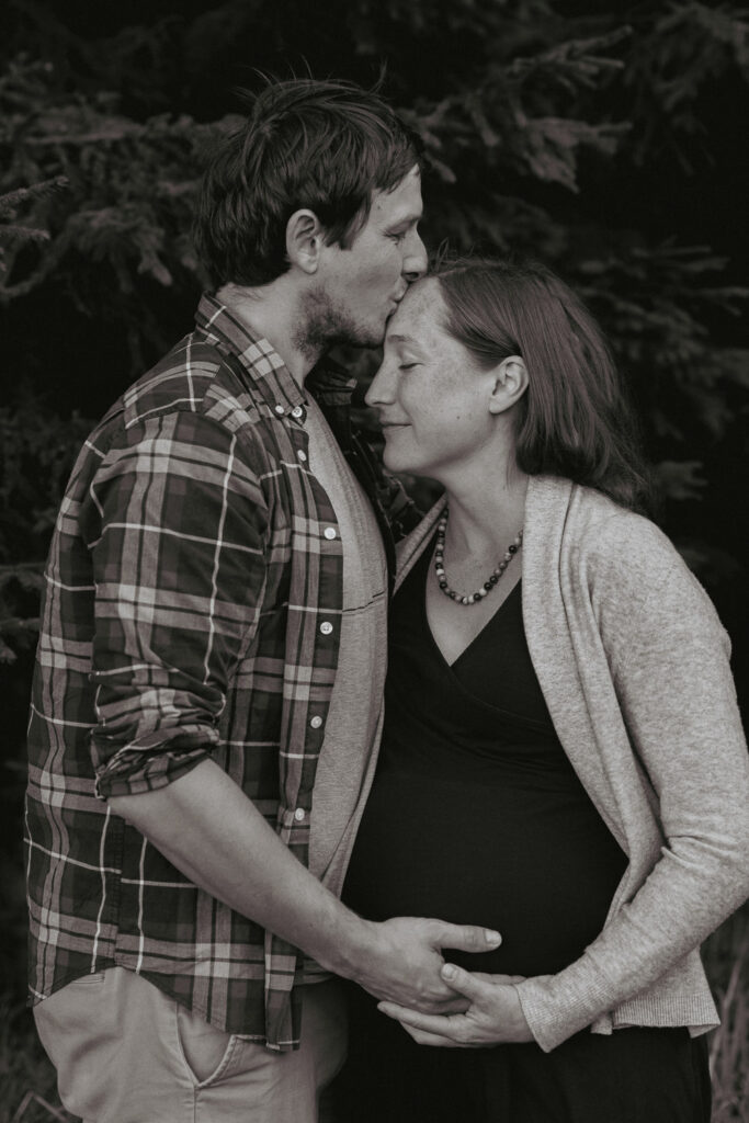 Ein Schwarz-Weiß-Bild. Ein Mann küsst seine schwangere Frau auf die Stirn.
