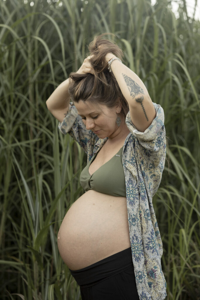 Schwangere Frau im hohen Gras. Sie trägt ein Bikinioberteil und eine offene Bluse.