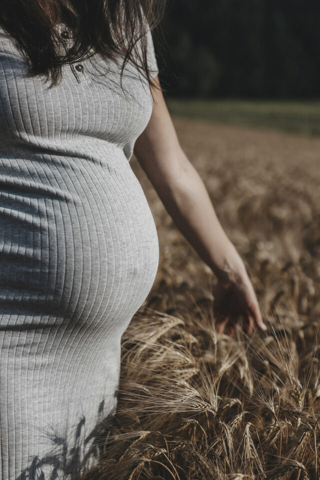 Ausschnitt einer schwangeren Frau in einem Getreidefeld. Mit einer Hand streicht sie über das Getreide.