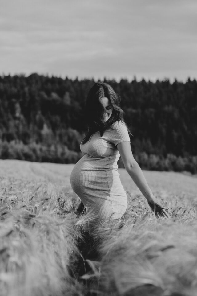 Eine schwangere Frau tanzt im Getreidefeld. Die Haare fallen ihr ins Gesicht. Das Bild ist in Schwarz-Weiß.