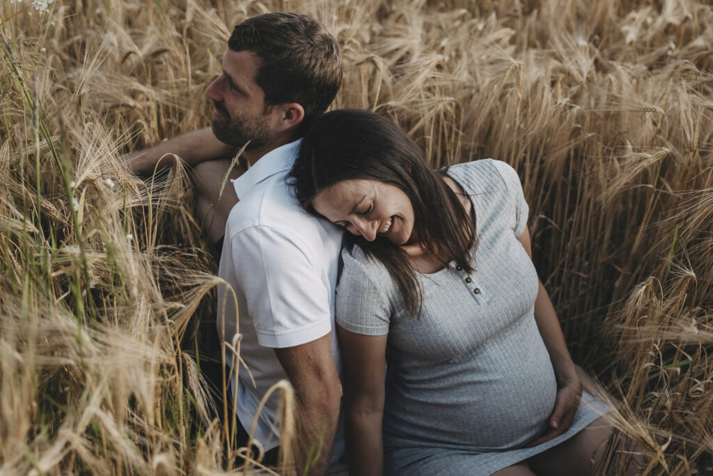 Ein Paar sitzt Rücken an Rücken im Getreidefeld. Beide lächeln. Die Frau ist schwanger und hält mit einer Hand ihren Bauch.