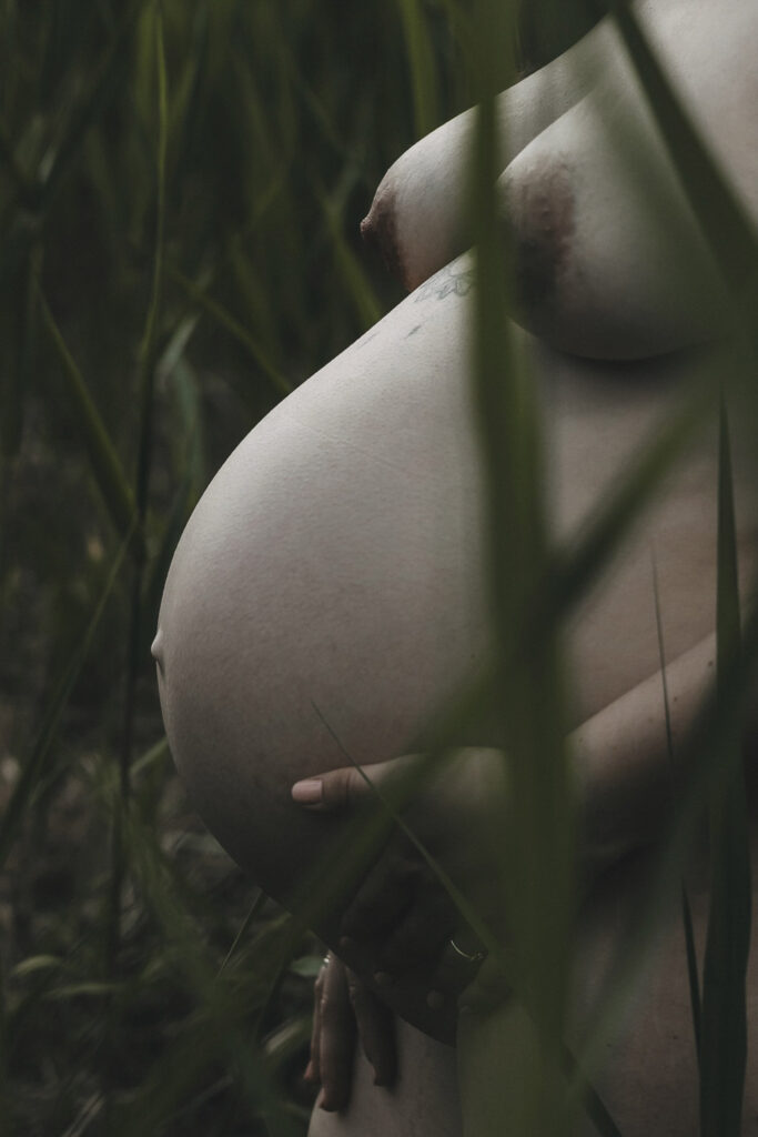 Seitenansicht einer nackten Frau. Man erkennt die Brüste und den Babybauch. Der Bauchnabel ragt heraus. Im Vordergrund und Hintergrund ist hohes, grünes Gras.