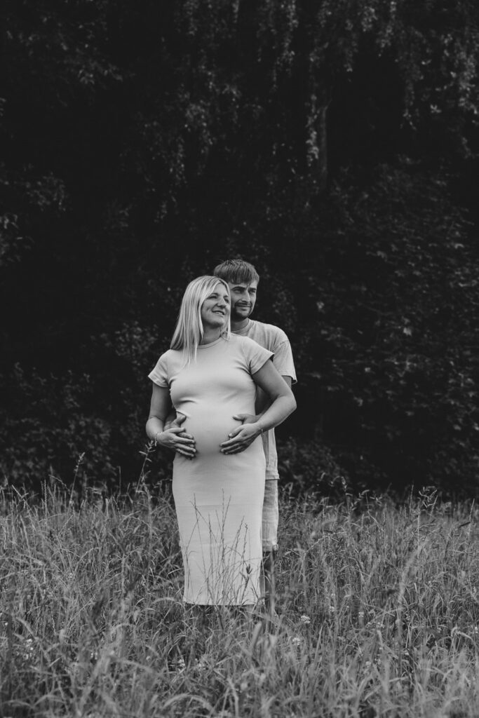 Schwarz-Weiß-Bild das eine schwanger Frau in einem Kleid zeigt, dahinter steht ihr Mann. Beide berühren den Babybauch. Es ist ein Ganzkörperportrait, beide Personen haben den Blick zur Seite gedreht.