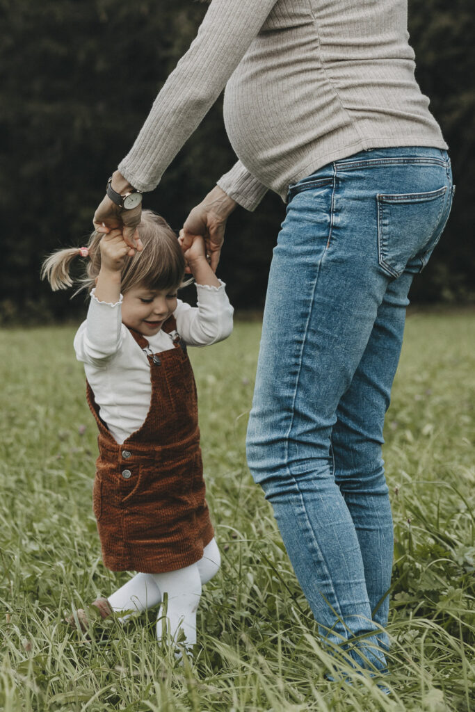 Tochter tanz mit ihrer schwangeren Mama im Kreis. Von der Frau sind nur die Beine erkennbar.