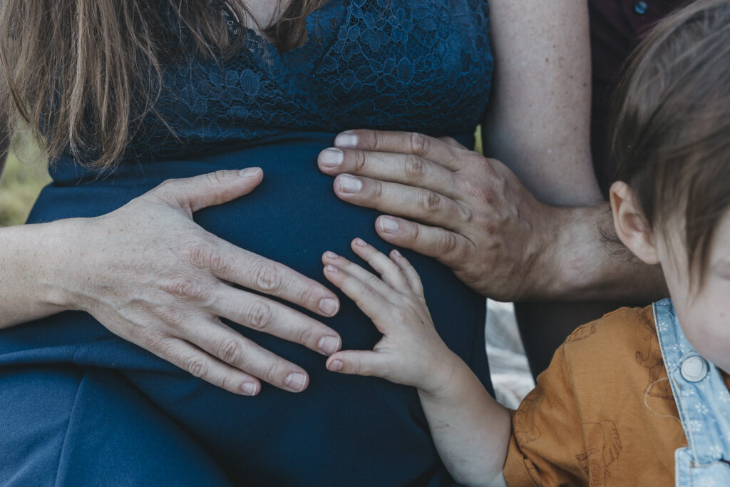 Die Hand einer Frau, eines Mannes und eines Kindes halten den Babybauch der Frau im blauen Kleid.