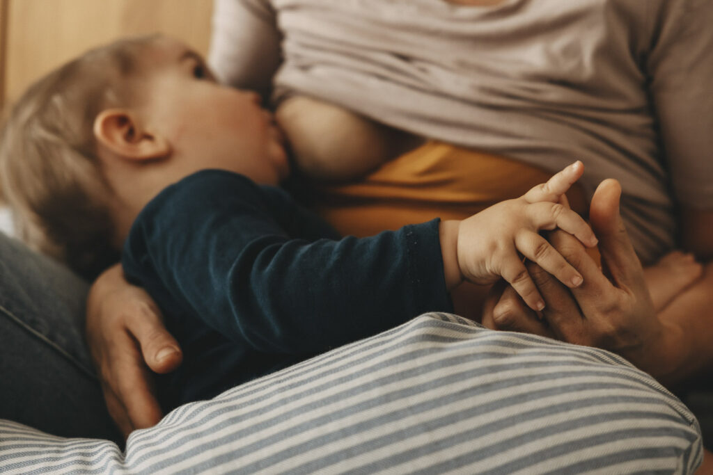 Mutter, die ihr einjähriges Kind stillt. Im Vordergrund sieht man die Hände der Beiden, die sich halten. Am Hintergrund der Blick des Babys zu seiner Mama.