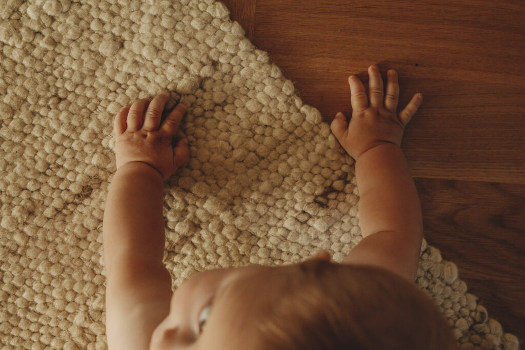 Baby, das sich mit den Händen zum ersten Mal aufstützt und versucht sich vorwärts zu wegen. Das Bild ist von oben fotografiert, man erkennt nur den Hinterkopf und die Hände.