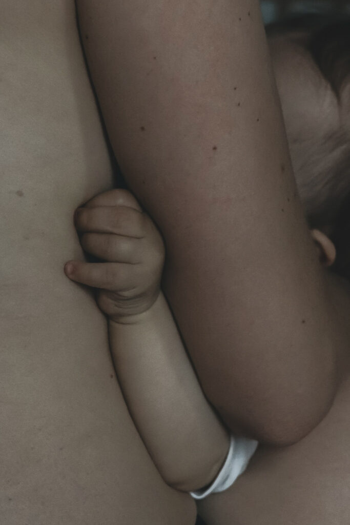 Ein nackter Babyarm und Hände, die sich am nackten Bauch der Mutter halten, während es gestillt wird.