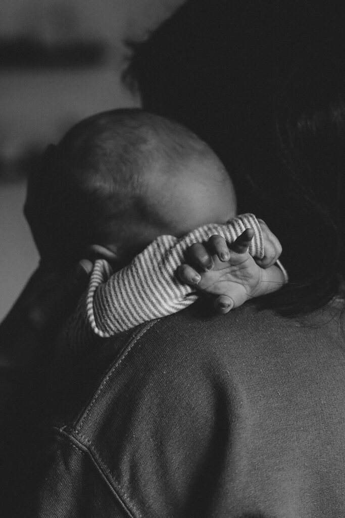 Baby auf Schulter der Mama. Den Blick versteckt es hinter seinen Händchen. Die Finger sind gestreckt. Schwarz-Weiß-Bild.