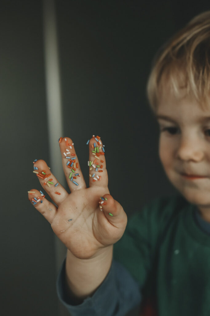 Junge, der seine Hand bestaunt. Er hat sie zuvor in bunte, süße Streusel getaucht.