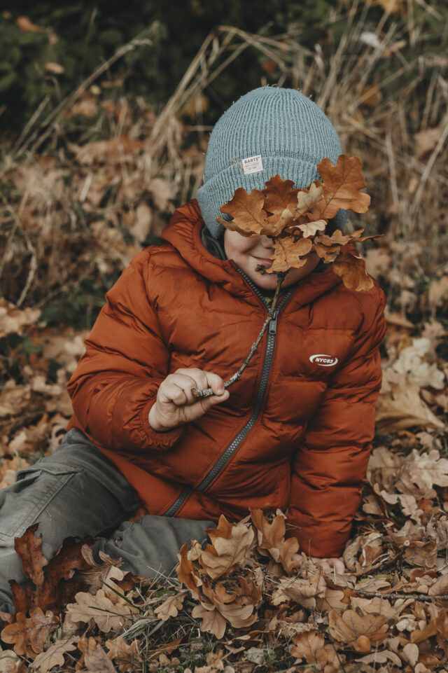 Kind in einem Blätterberg, das Gesicht ist hinter einem Ast mit Blättern versteckt.