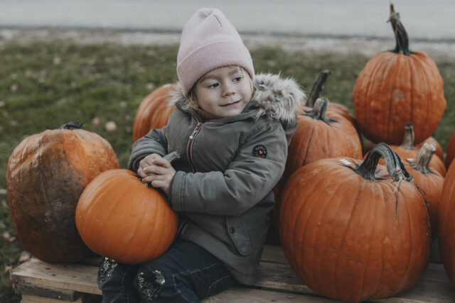 Mädchen in einer dicken Jacke und mit einer Haube auf dem Kopf umringt von organgen Kürbissen.