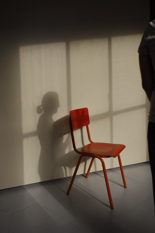 Ein roter Stuhl dahinter ein Schatten einer Frau mit Zopf und der Schatten eines Fensters.