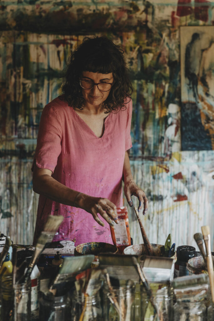 Malerin in einem rose Kleid. Im Vordergrund stehen Pinseln und Farben auf einem Tisch. Im Hintergrund erkennt man das bunte Atelier und ein Bild, das gerade gemalt wird.