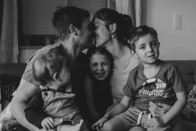 Familienbild mit Eltern, die sich küssen und drei Kinder auf ihren Schoß haben. Schwarz-Weiß-Bild.