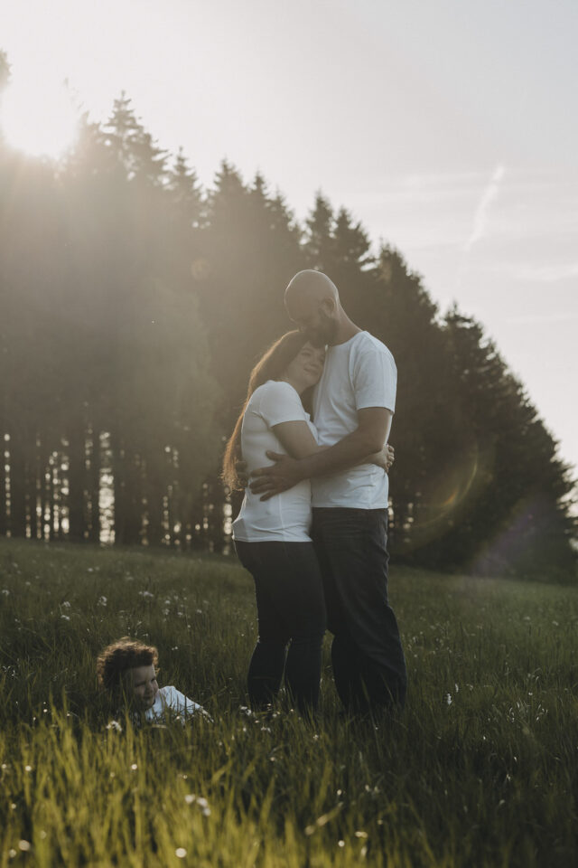 Paar, das in einer Wiese am Waldrand steht und sich umarmt. Am Boden liegt ein Dreijähriger und turnt herum.