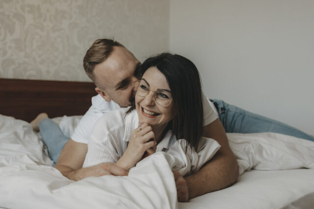 Ein junges Pärchen, dass im Bett liegt. Der Mann flüstert der Frau etwas ins Ohr, dass sie zum Lächeln bringt.