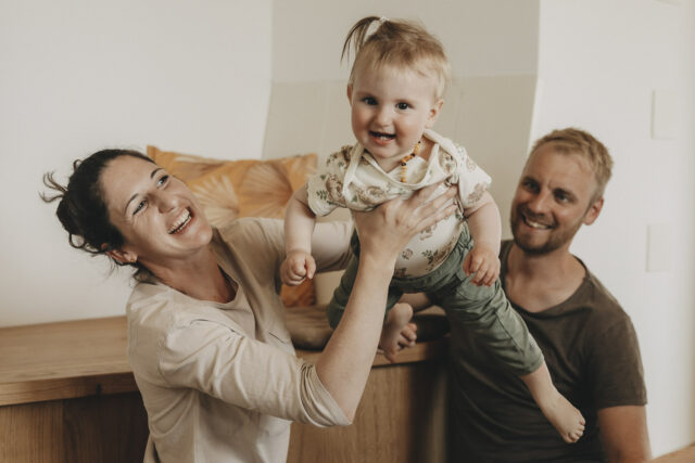 Mama hebt ein lachendes Kind hoch, es fliegt. Der Papa im Hintergrund strahlt.