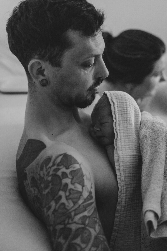 Neugeborenes liegt zugedeckt, Haut auf Haut, an der Brust seines Vaters. Schwarz-Weiß-Bild.