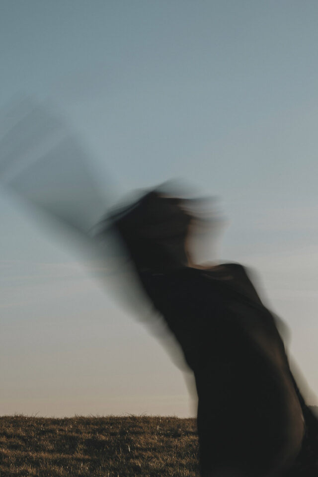 Tanzende Frau mit Bewegungsunschärfe. Im Hintergrund ist der Himmel zu erkennen, bei Sonnenuntergang.