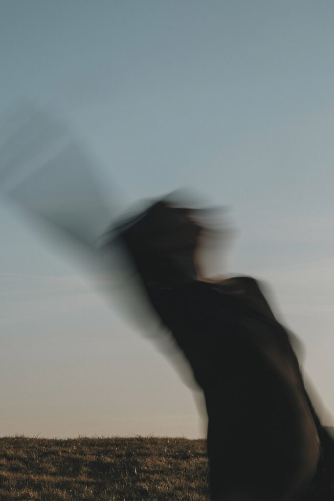 Tanzende Frau mit Bewegungsunschärfe. Im Hintergrund ist der Himmel zu erkennen, bei Sonnenuntergang.