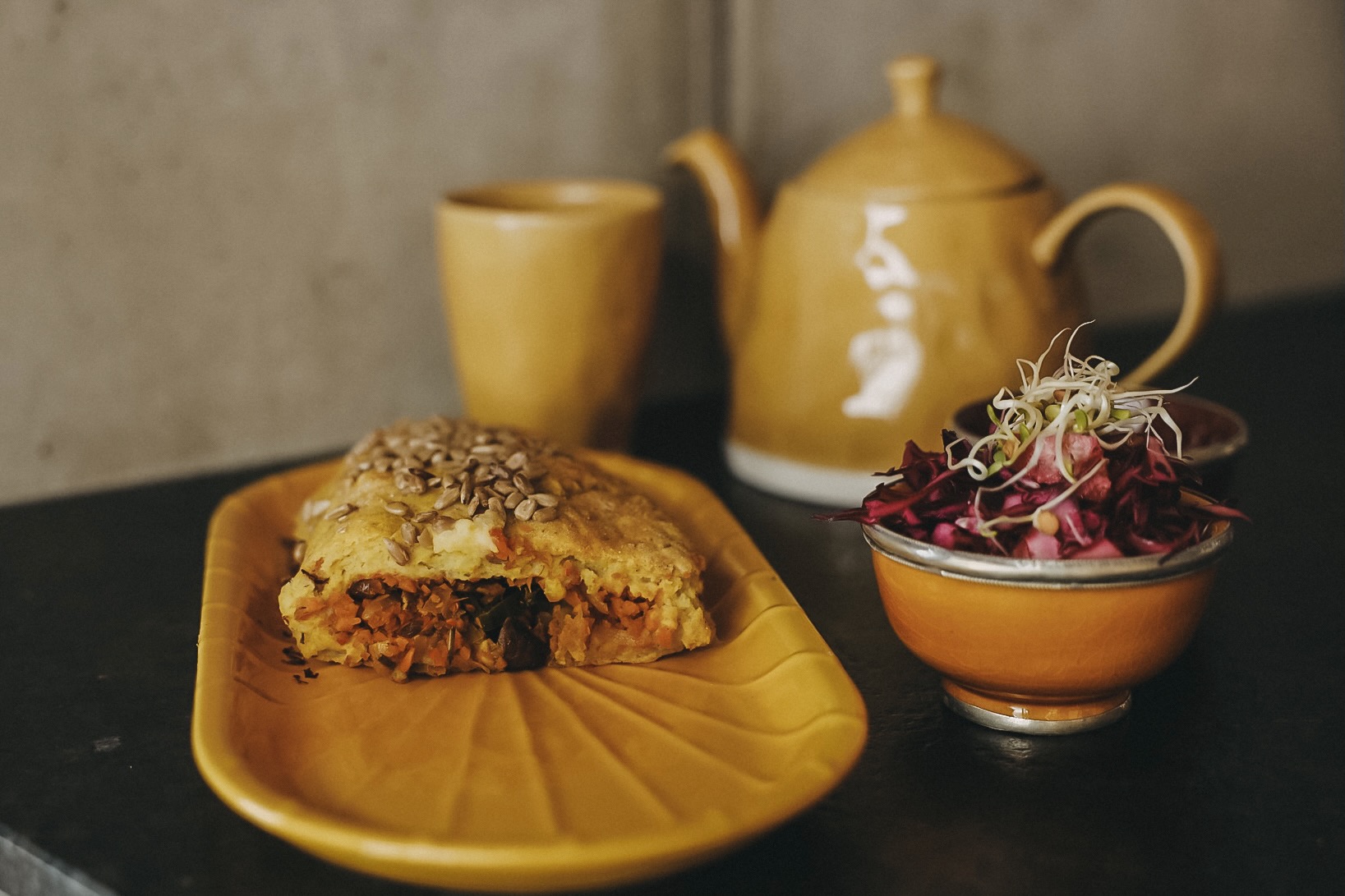 Kürbisstrudel auf einem gelben Teller, daneben ein Rote-Rüben-Salat in einer orangen Schüssel. Im Hintergrund erkennt man eine gelbe Kanne Tee und eine Tasse dazu.