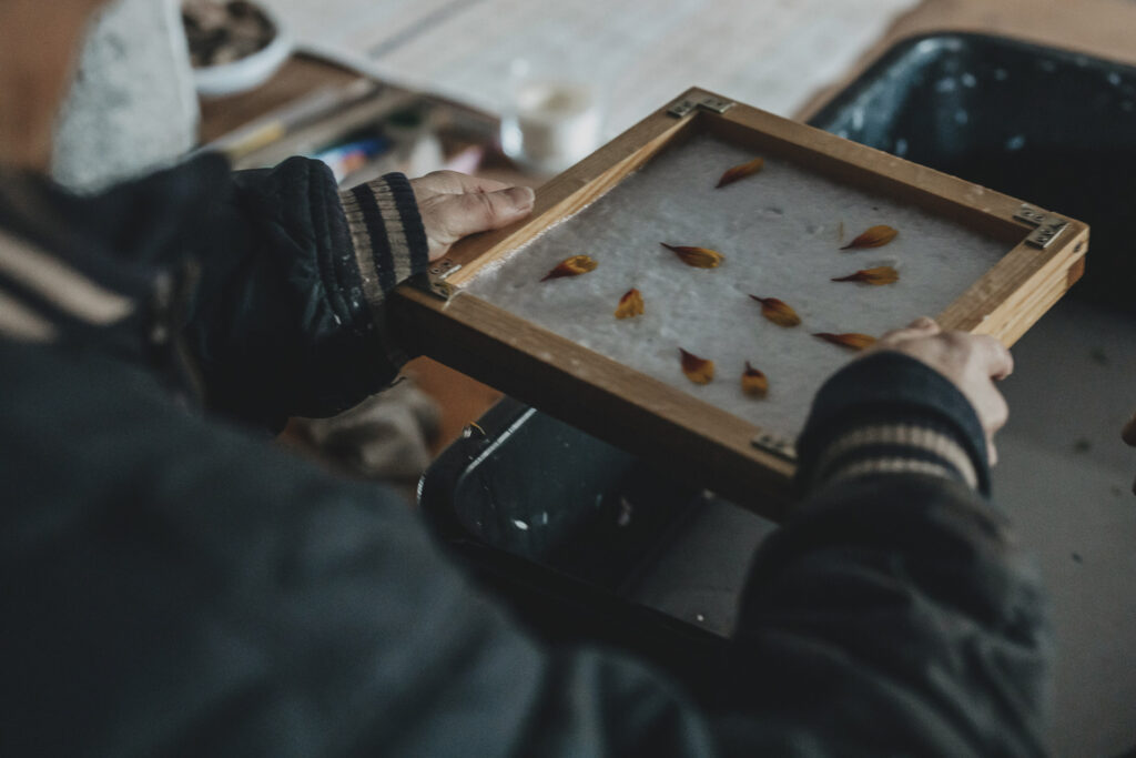Hände die den Rahmen und das geschöpfte Papier mit Blüten halten.