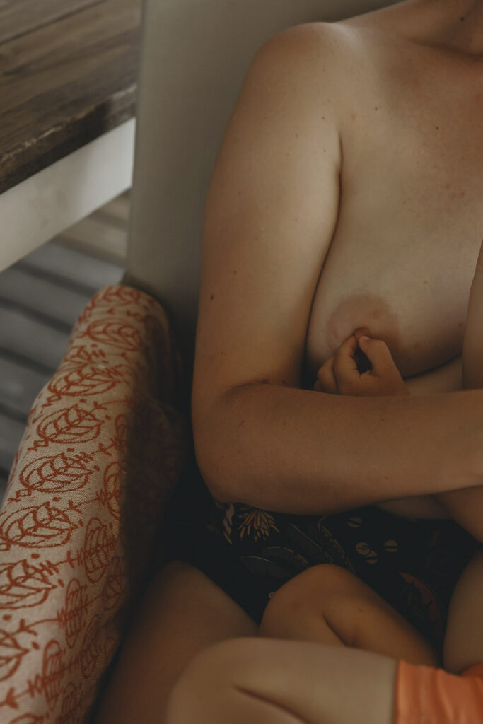 Eine nackte Frau - zu erkennen ist ein Teil ihres Armen und ihre Brust. Ein Kind berührt sanft ihre Brustwarze, zur Beruhigung.