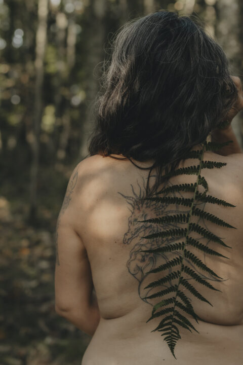 Nackte Frau von Hinten. Ihr Rücken ist teilweise bedeckt mit einem großen, grünen Farn.