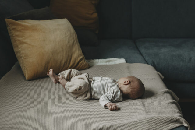 Kleines Baby liegt auf der Couch. Die Füßchen in die Höhe gestreckt, den Blick zur Seite gerichtet, nicht erkennbar.