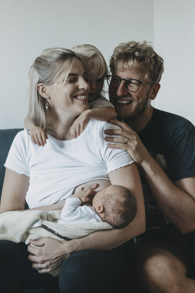 Familienbild mit Mama, Papa, einem Kind und einem Baby. Alle umarmen sich und lachen sich gegenseitig an.