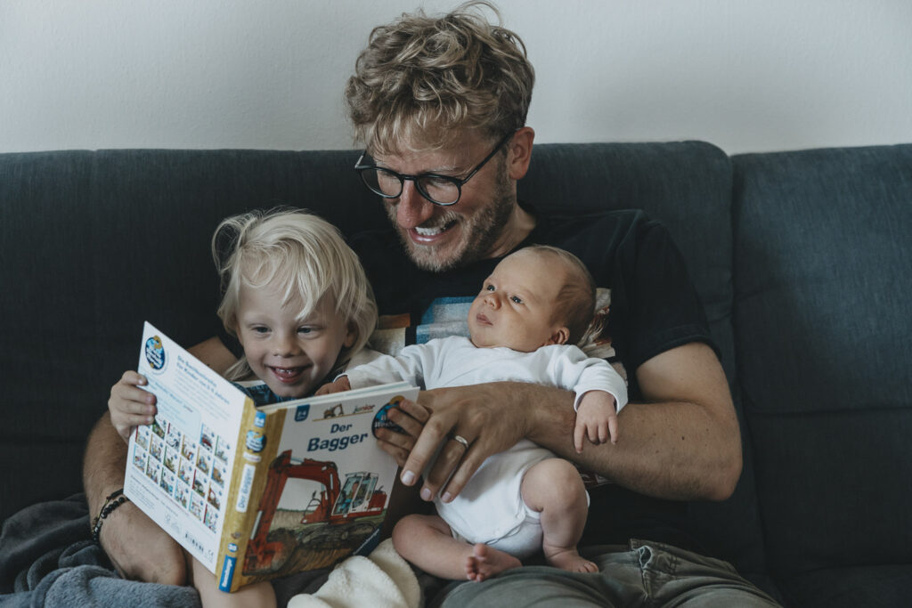 Vater mit zwei Kindern auf dem Schoß, sie schauen sich gemeinsam ein Buch an.