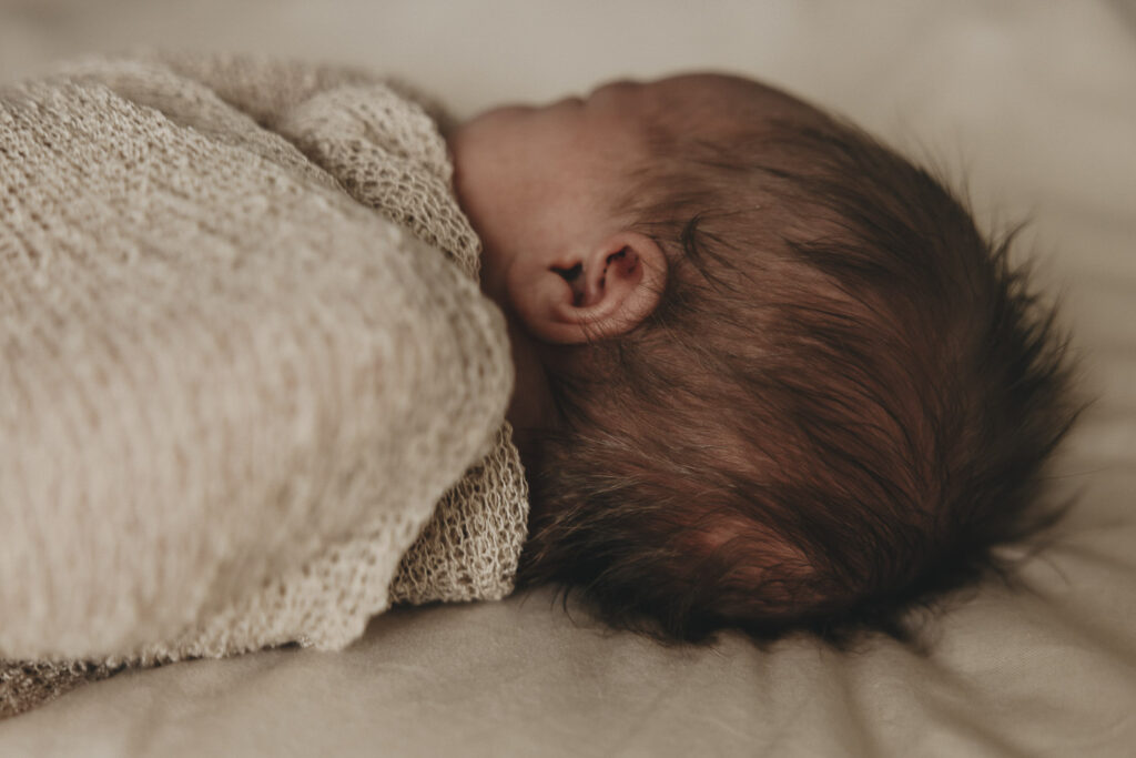 Baby eingewickelt in ein weiches Tuch. Zu erkennen ist das Köpfchen von Hinten mit vielen, dunklen Haaren.