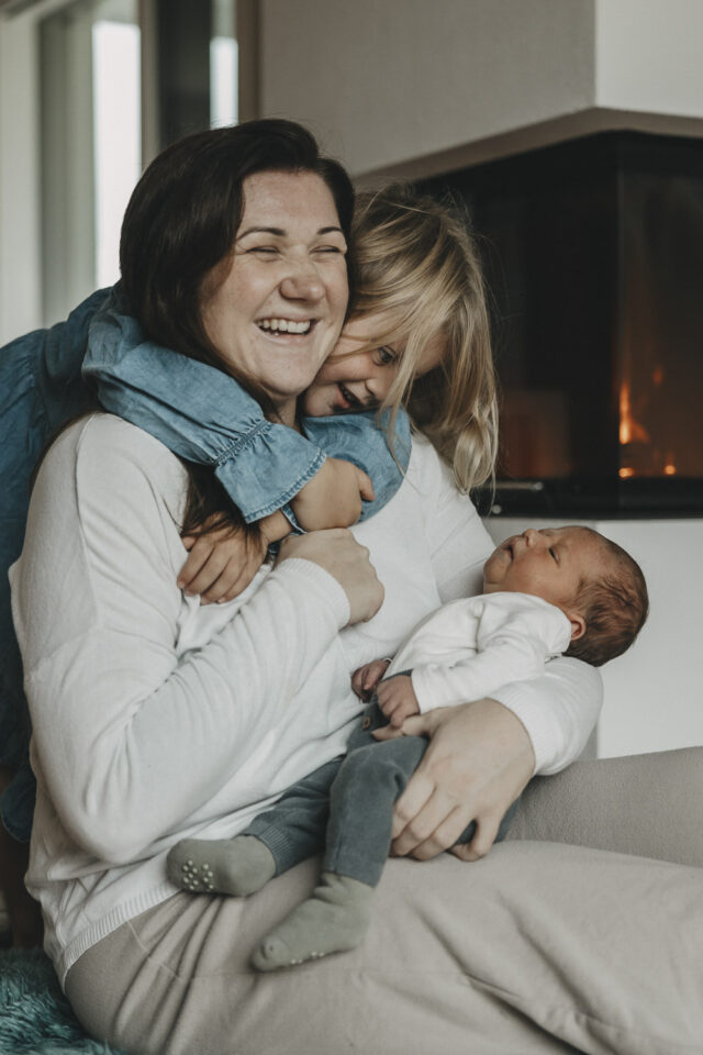 Mutter hält ihr Neugeborenes in den Armen. Das Geschwisterkind umarmt sich gleichzeitig ganz fest. Die Mama strahlt bis über beide Ohren.