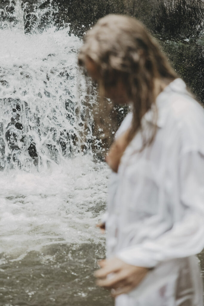 Frau mit naßen Haaren und weißer Bluse unscharf im Vordergrund. Dahinter ein reißender Fluss.